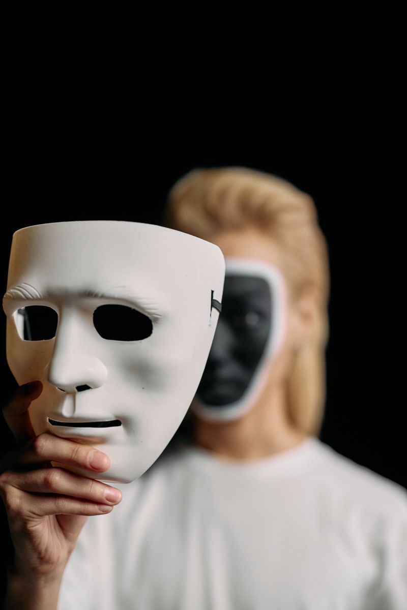 A person holds a white mask in a studio with face obscured by creative black and white makeup.